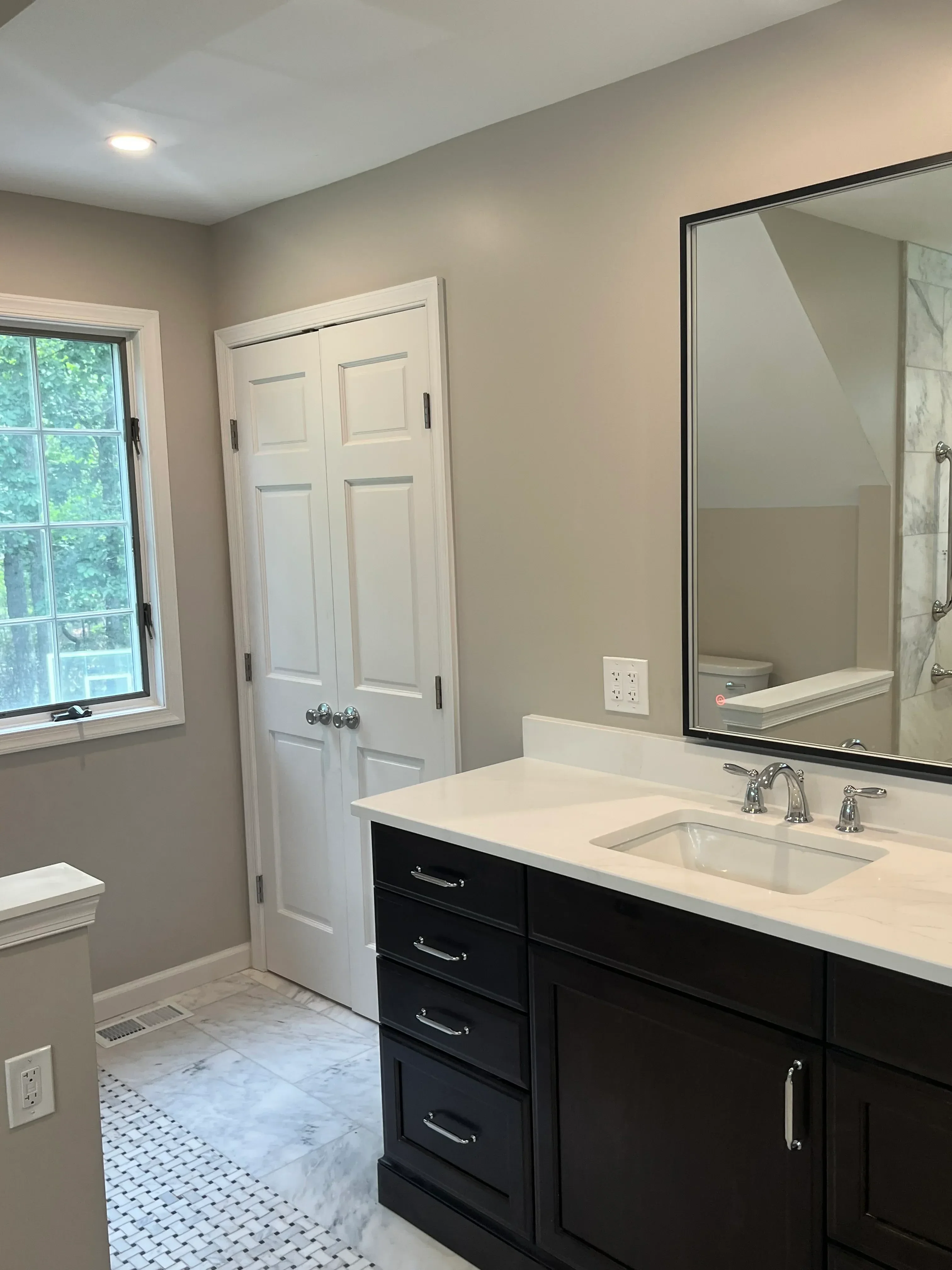 White bathroom vanity, marble tile shower, framed mirror, chrome fixtures, and black cabinet hardware