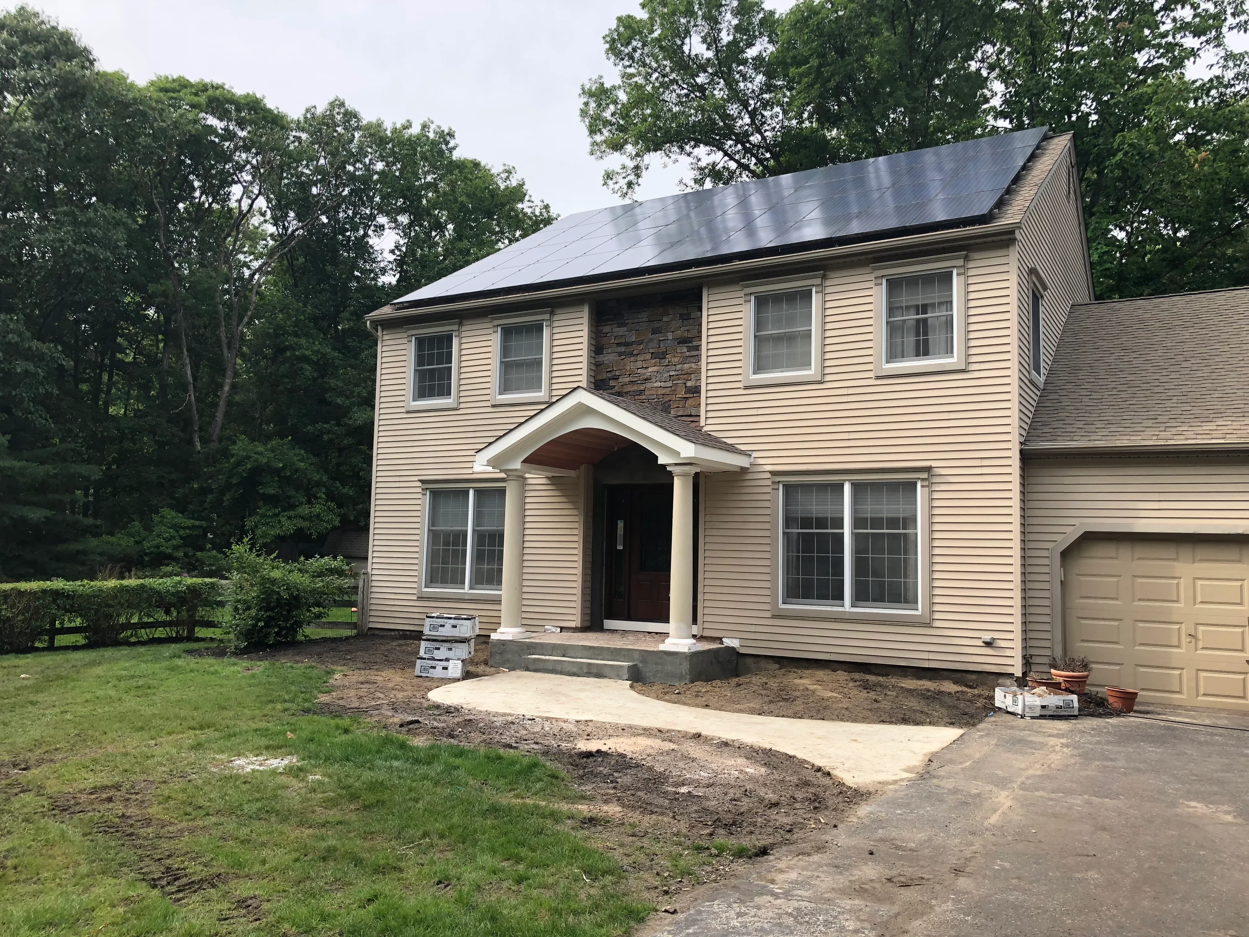 Two-story home with tan vinyl siding, dark architectural shingle roof, stone chimney, and new front portico with columns