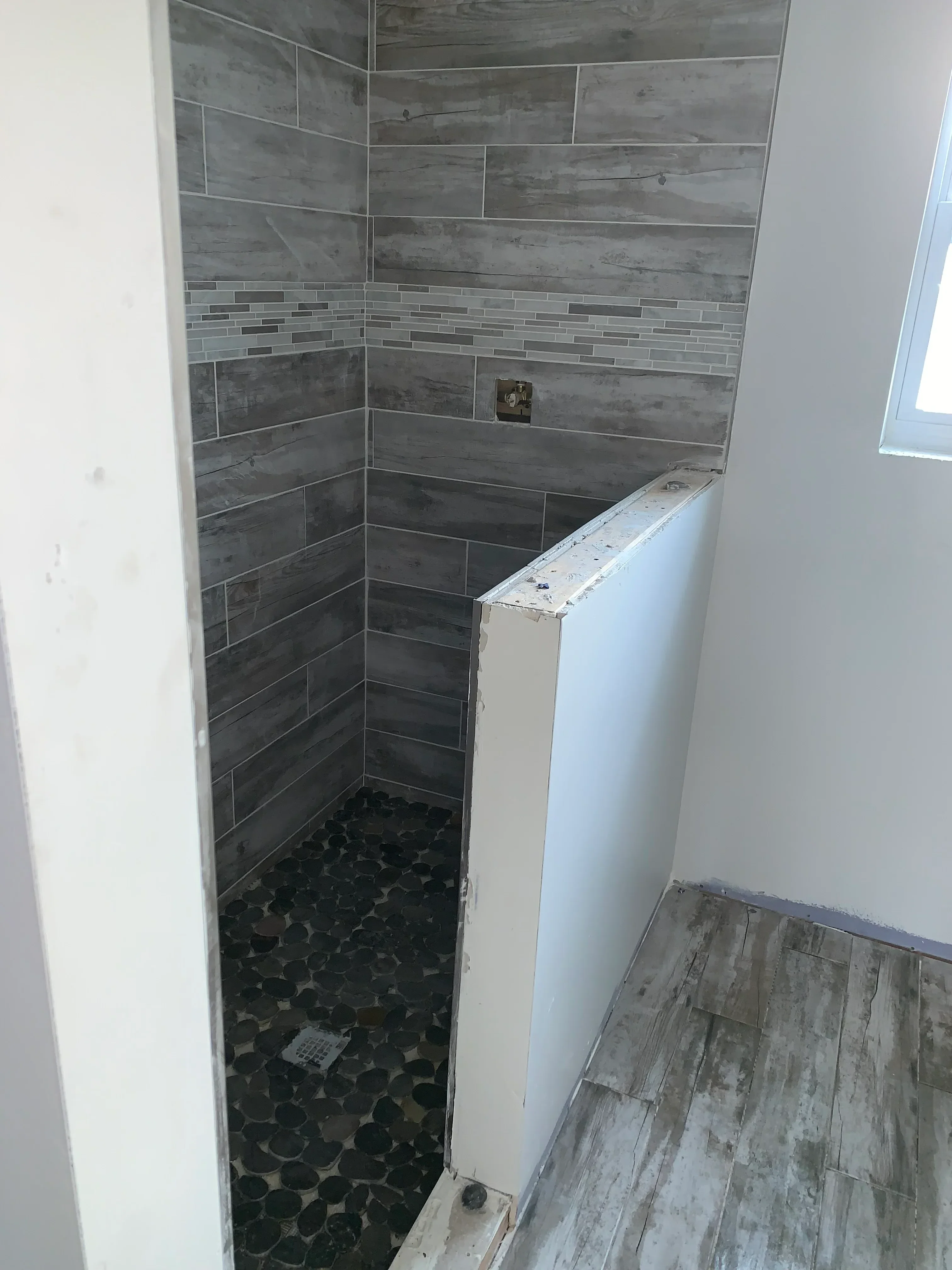 Gray wood-look plank tile flooring with linear accent strips and dark pebble tile accent wall in bathroom